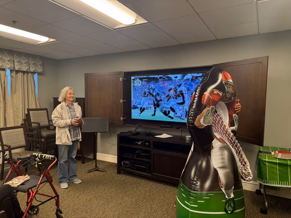 A senior woman holding a football stands in a room with a television showing a football game. There is an inflatable football player figure in front of the TV, and a walker is visible in the foreground. The room has chairs stacked against the wall and a table with a green football-themed tablecloth.