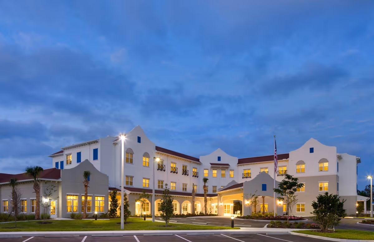Exterior front view of a three-story senior living facility at dusk with lit windows and a landscaped entrance.