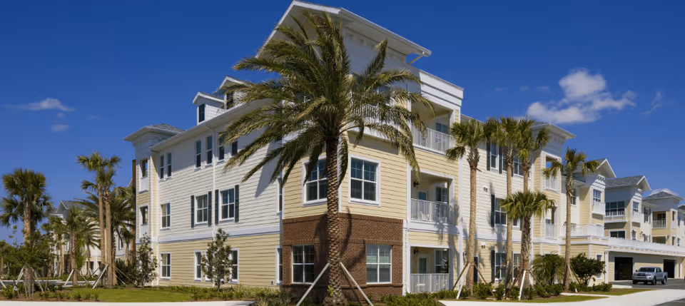 Front exterior of a three-story senior living building with balconies, palm trees, and a bright blue sky.