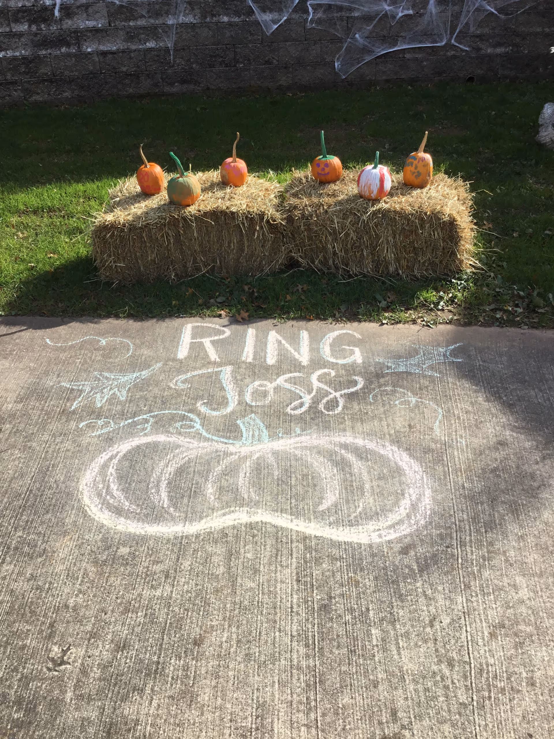 Painted pumpkins sit on hay bales in front of a stone wall with spiderweb decorations and a sidewalk chalk 'Ring Toss' drawing.
