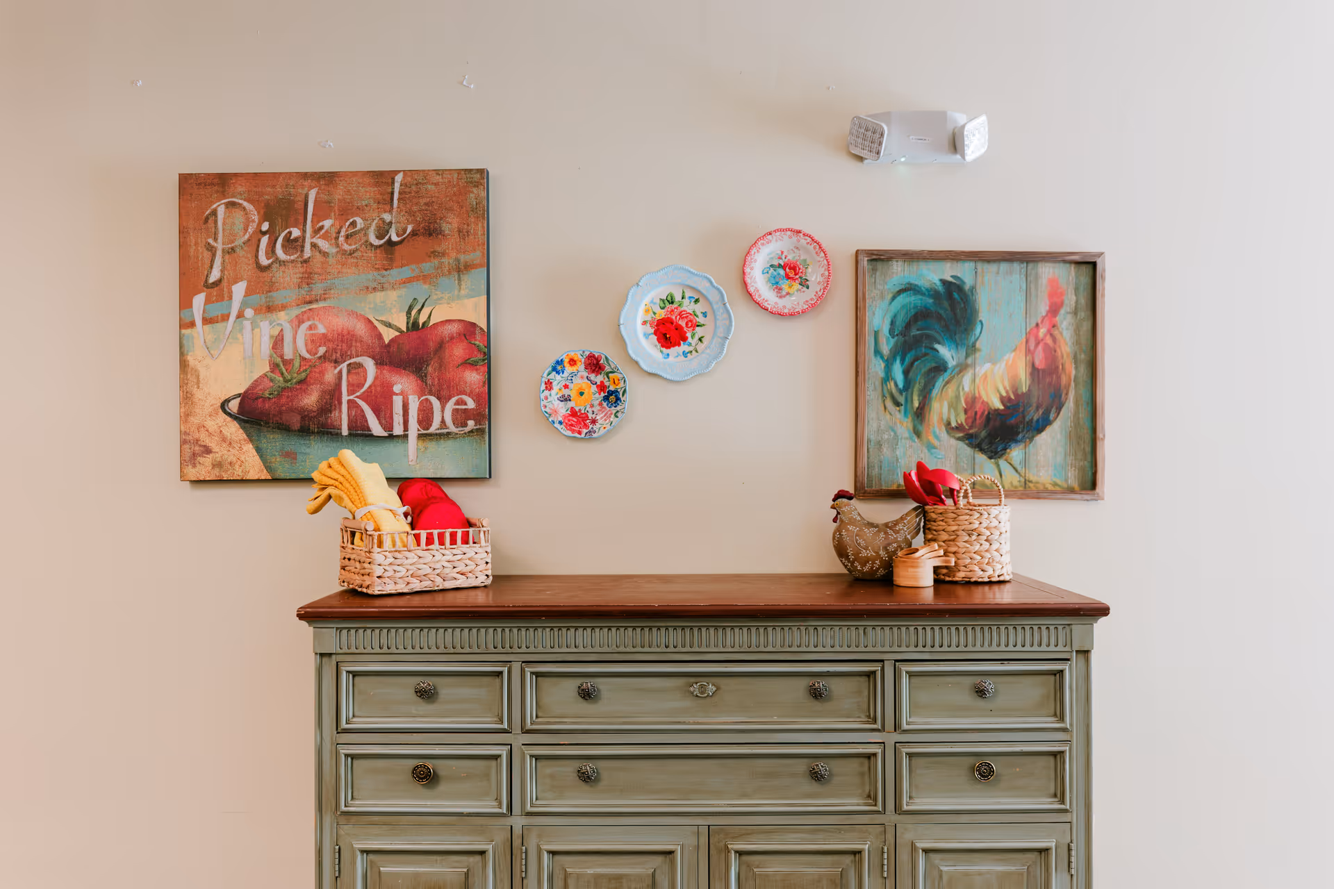 A decorative green wooden sideboard with a dark wooden top against a beige wall. On the wall above the sideboard are two framed pictures, one with tomatoes and the text 'Picked Vine Ripe' and the other with a colorful rooster. Between the pictures are three decorative floral plates. On the sideboard are two woven baskets containing folded cloths and a small ceramic hen figurine.