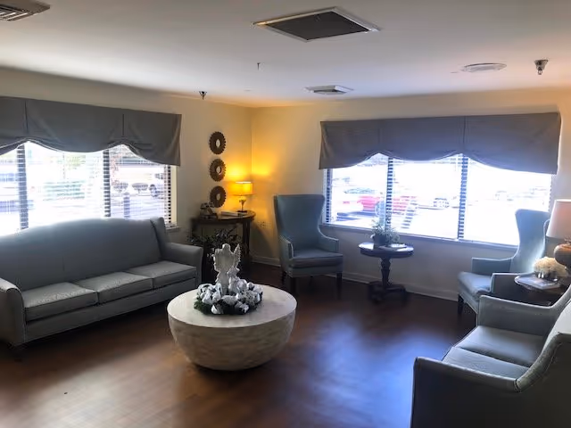 A well-lit living room with large windows covered by valance curtains. The room features a gray sofa, two gray armchairs, a round coffee table with a decorative centerpiece, a small side table with a lamp, and wooden flooring. The walls are light-colored, and there are decorative wall hangings near the corner.