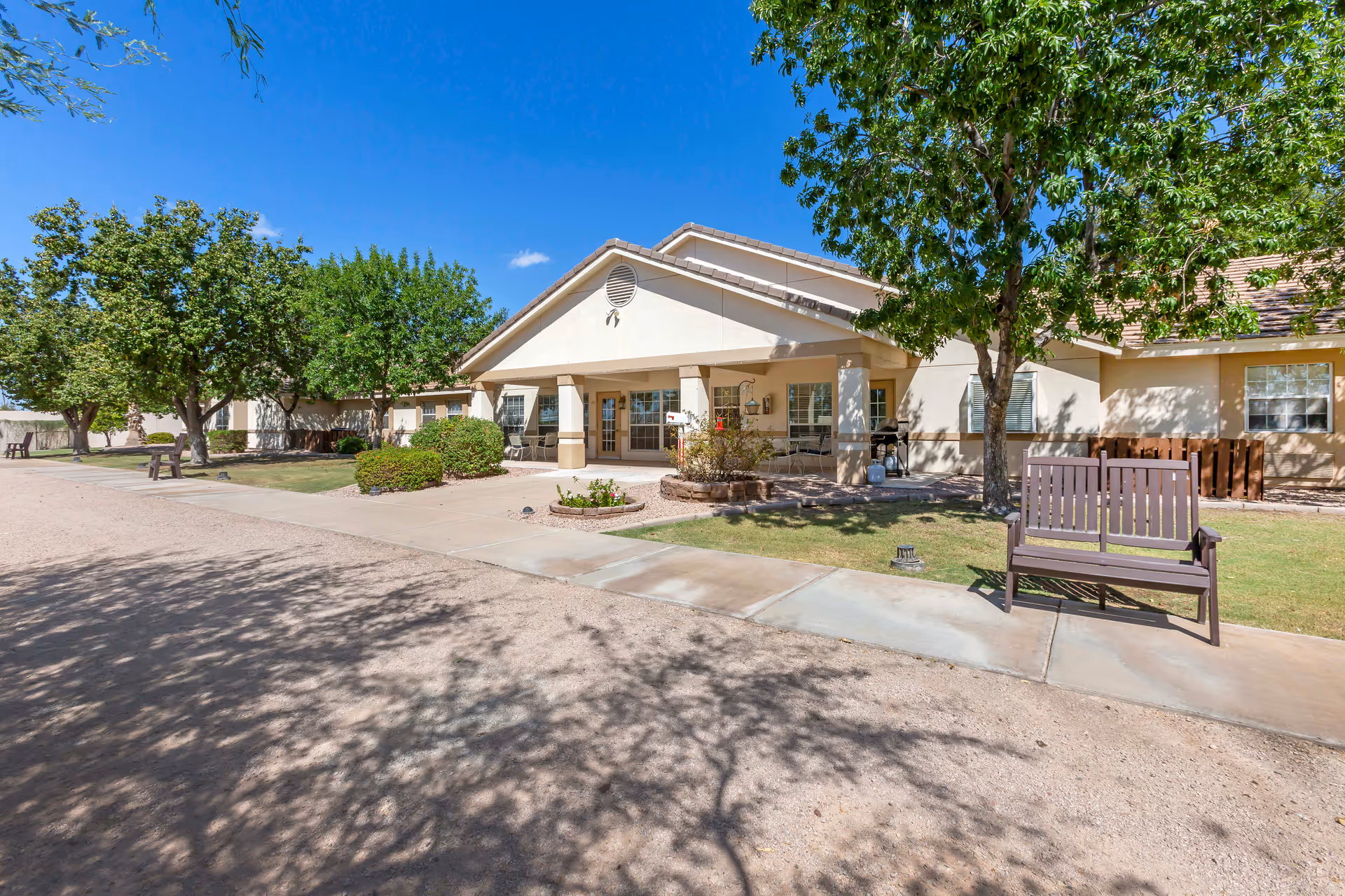 Exterior view of a single-story senior living facility building with a covered porch, surrounded by green trees and bushes under a clear blue sky. There is a bench on the sidewalk in front of the building.