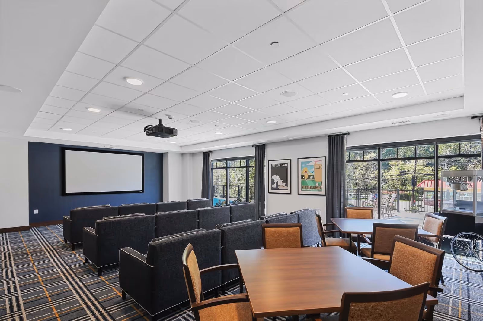 A modern community room with rows of dark gray armchairs facing a large blank projector screen mounted on a navy blue accent wall. The room has a patterned carpet with gray, black, and orange stripes, and several wooden tables with brown cushioned chairs. Large windows with dark curtains let in natural light and offer a view of an outdoor patio area with chairs and greenery. Two framed Pittsburgh-themed posters hang on the wall between the windows.