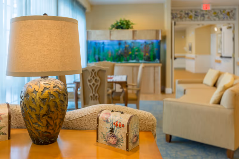 Well-lit common room with a decorative table lamp and small decorative chests in the foreground, seating and an aquarium visible in the background.