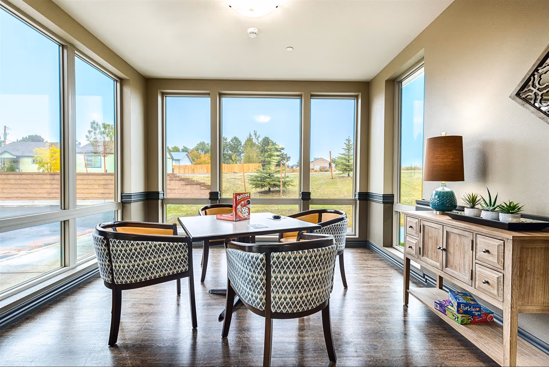 A bright room with large windows showing an outdoor view of trees and houses. Inside, there is a square table with four patterned chairs around it. On the table, there is a Yahtzee game box and some papers. To the right, there is a wooden sideboard with a lamp, decorative plants, and board games on the lower shelf. The floor is wooden, and the walls are painted light beige.