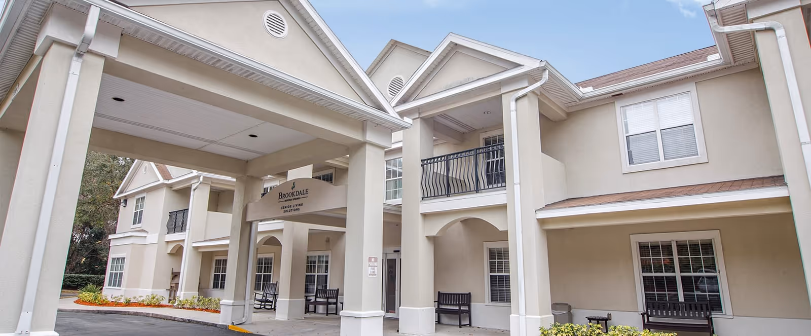 Exterior view of the Brookdale Wekiwa Springs senior living facility showing a beige two-story building with a covered entrance supported by columns, benches near the entrance, and windows with white frames.