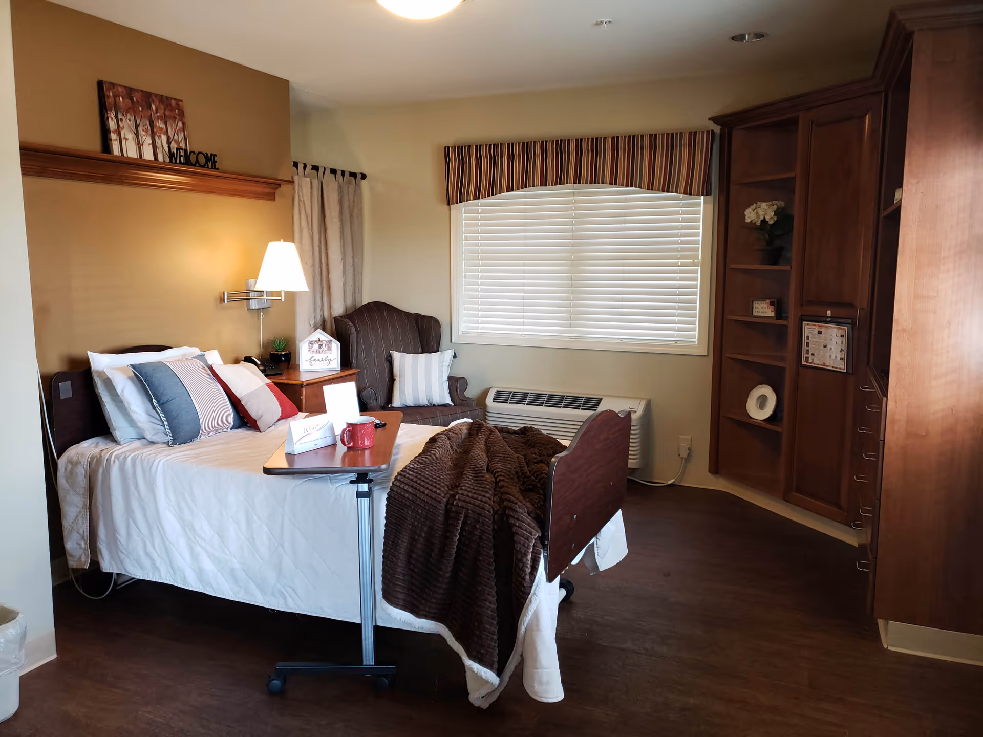 A cozy bedroom in a senior living facility with a single bed covered in white bedding and multiple pillows. A brown blanket is draped over the foot of the bed. Next to the bed is a small wooden nightstand with a lamp, a small plant, and a decorative sign. A comfortable armchair with a striped pillow is positioned near a window with closed blinds and a striped valance. There is a wooden built-in shelving unit and cabinetry on the right side of the room. The floor is a dark wood laminate.