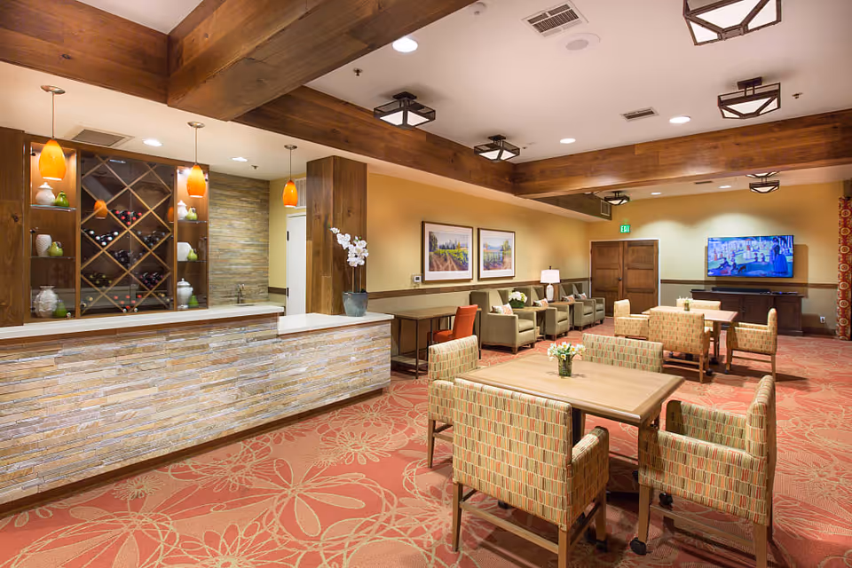 A cozy common area in a senior living facility featuring wooden beams on the ceiling, a stone and wood reception or bar counter with wine bottles and decorative items on shelves, several tables with patterned chairs, armchairs along the wall, framed landscape paintings, a flat-screen TV mounted on the wall, and warm lighting.