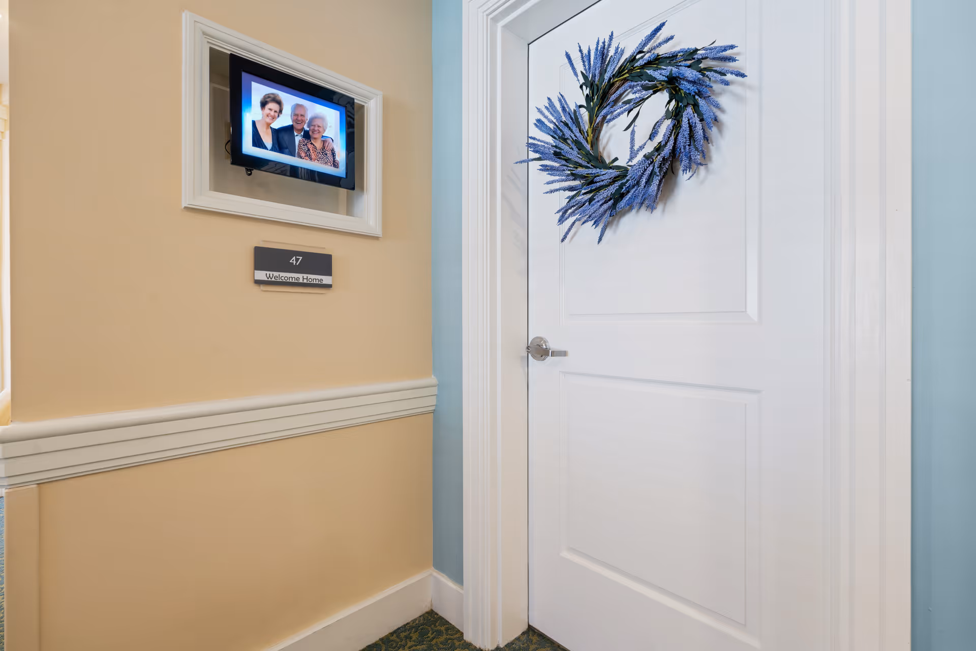 A white door decorated with a blue floral wreath in a hallway with beige and light blue walls. Next to the door is a framed digital photo of three elderly people and a sign below it that reads '47 Welcome Home'.