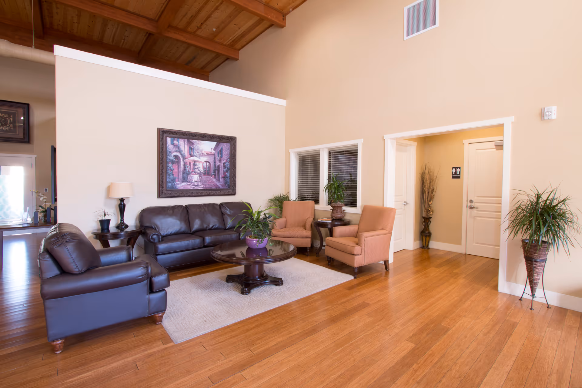 A spacious communal living room with leather sofas, armchairs, a coffee table, plants, and wood floors under a high wooden ceiling.