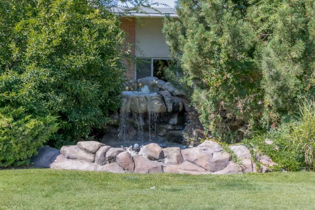 A small outdoor water feature with rocks and a gentle waterfall surrounded by green bushes and trees, with a building partially visible in the background.