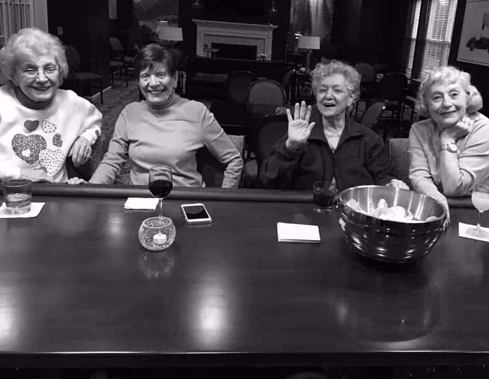 Four elderly women sitting at a bar counter in a senior living facility, smiling and enjoying drinks. One woman is waving at the camera. There is a large bowl of snacks on the counter along with glasses and a candle holder.