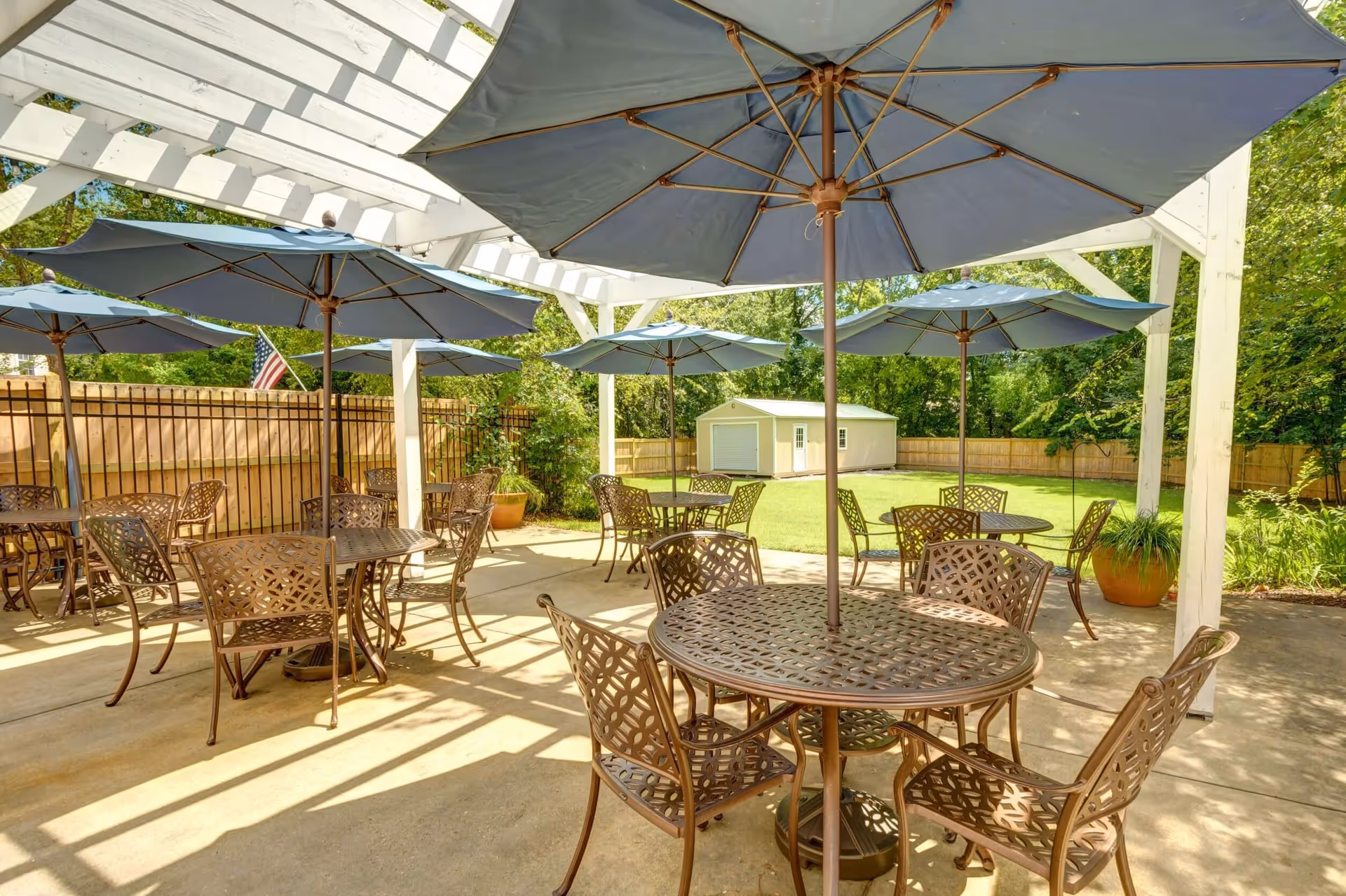 Outdoor patio area with multiple round metal tables and matching chairs, each table shaded by a large blue umbrella. The patio is covered by a white pergola, and there is a fenced grassy yard with a small shed in the background surrounded by trees and plants.