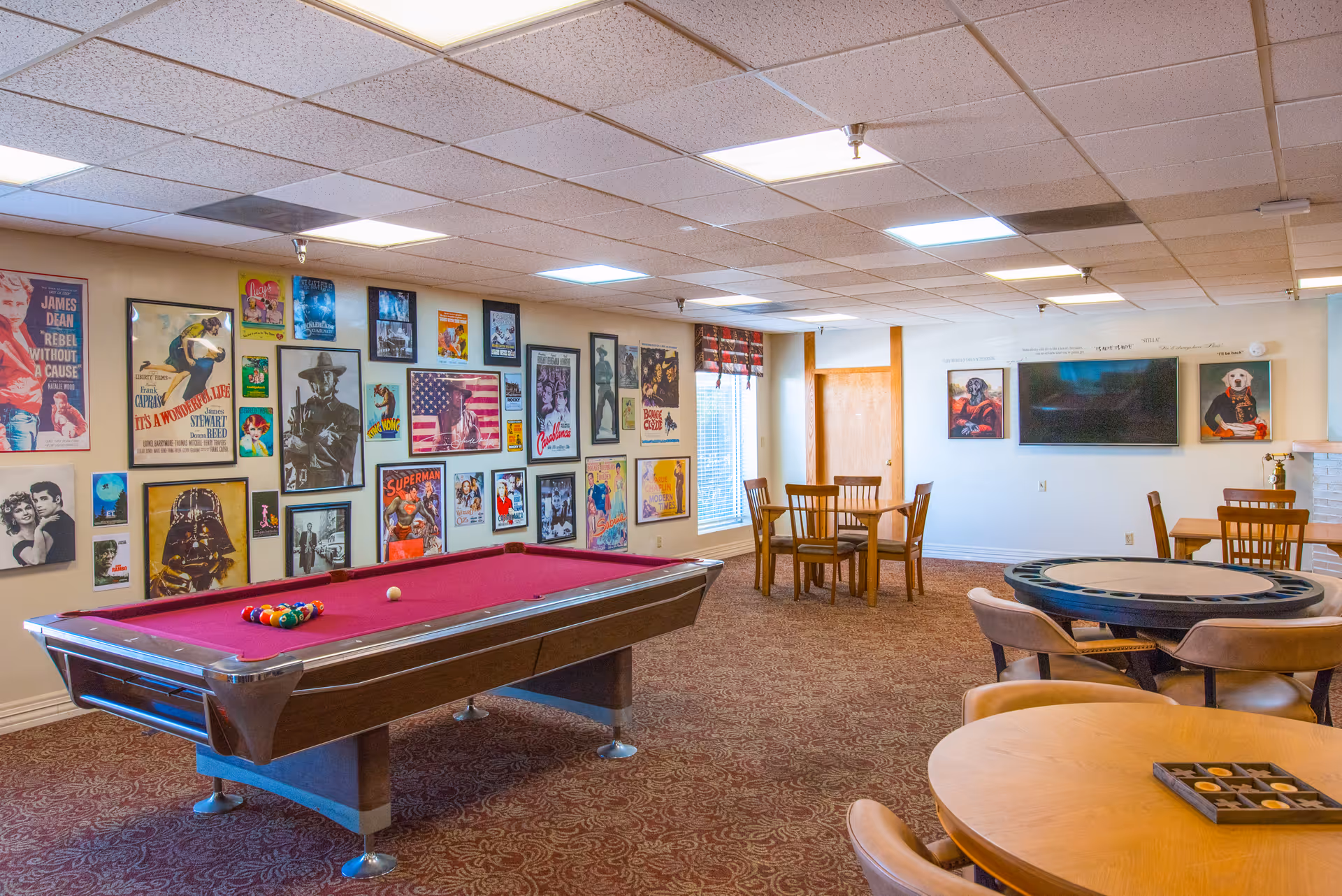 A recreational room with a red pool table in the center, surrounded by several wooden tables and chairs. The walls are decorated with numerous colorful vintage movie and pop culture posters. The room has a carpeted floor and a drop ceiling with fluorescent lighting.