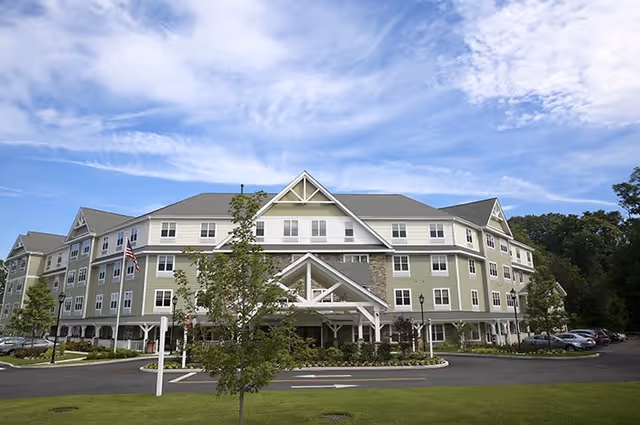 Large multi-story senior living facility with a covered entrance, landscaped grounds, and parked cars under a blue sky.