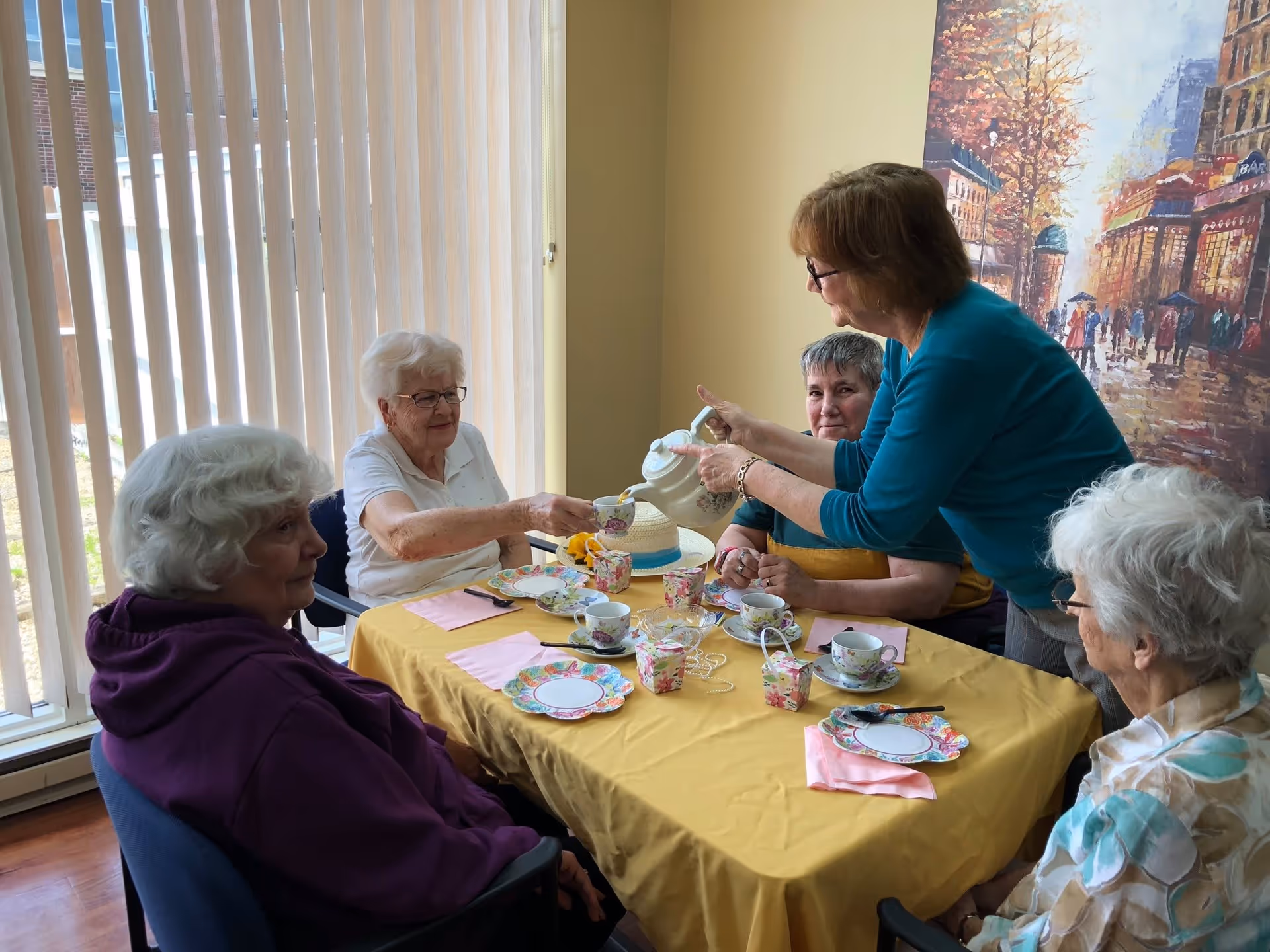 A group of elderly women sitting around a table with a yellow tablecloth, having tea. One woman is pouring tea from a teapot into a cup held by another woman. The table is set with floral-patterned teacups, saucers, plates, and pink napkins. The room has vertical blinds covering a window and a large colorful painting on the wall.