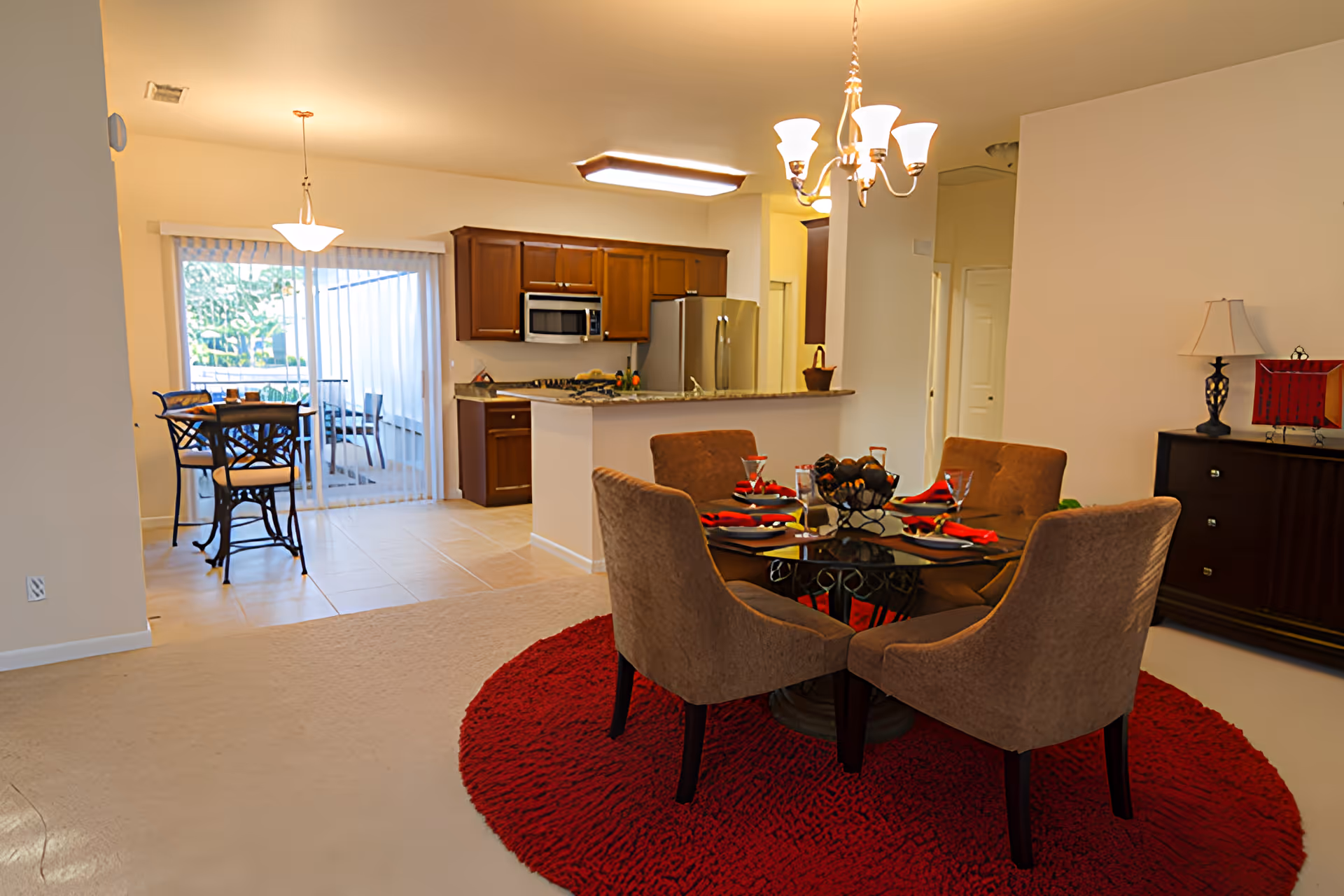Interior view of a senior living facility dining area and kitchen at Paradise Valley Estates. The dining area features a round glass table with four brown upholstered chairs on a red circular rug. The kitchen has wooden cabinets, a stainless steel refrigerator, microwave, and a countertop with a sink. In the background, there is a small breakfast nook with a table and chairs near sliding glass doors that lead to an outdoor space.