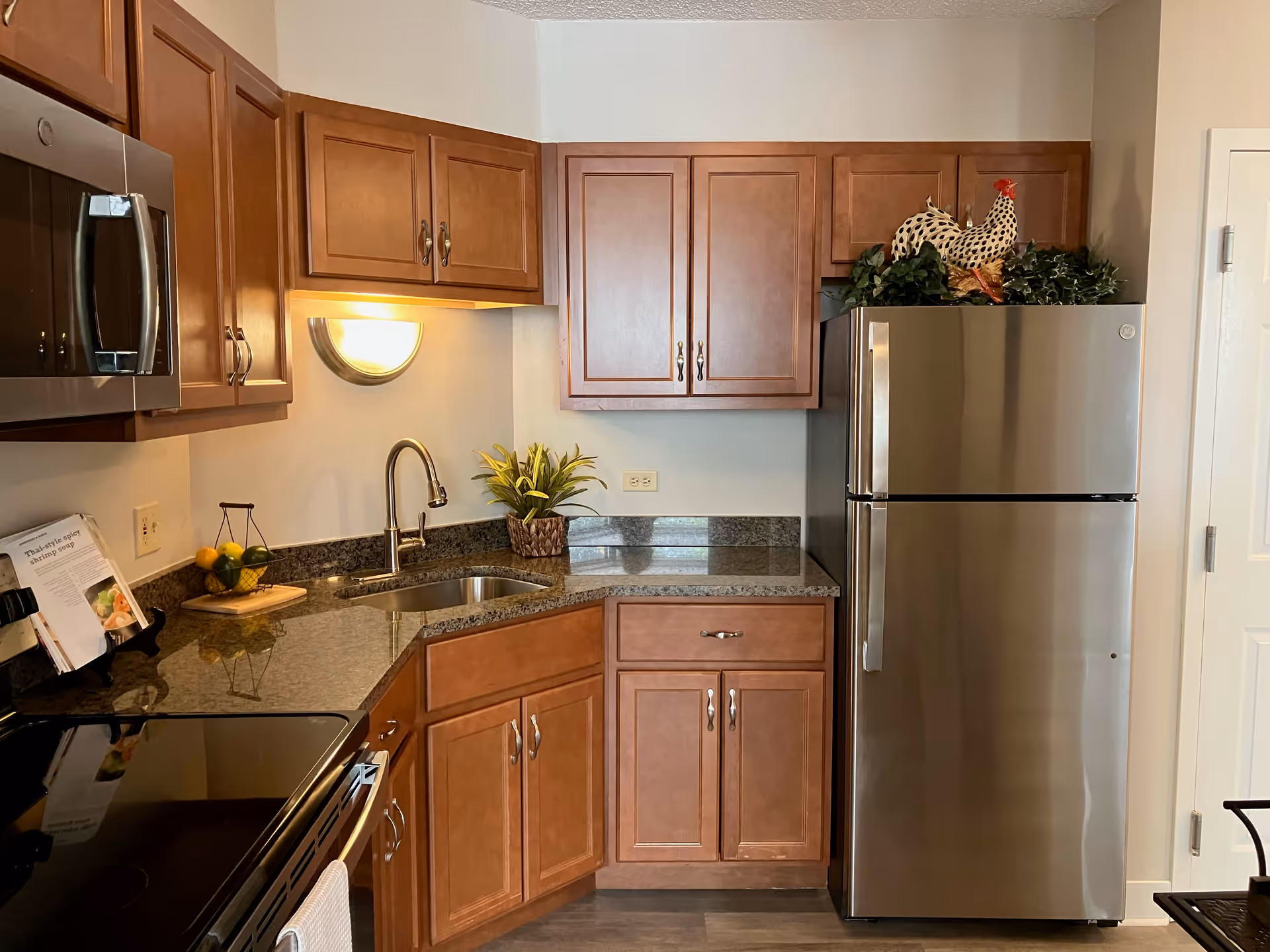 A modern kitchen with wooden cabinets, a stainless steel refrigerator, a built-in microwave, an electric stove, a granite countertop with a sink, a wall-mounted light fixture, a small plant, and a decorative rooster on top of the refrigerator.