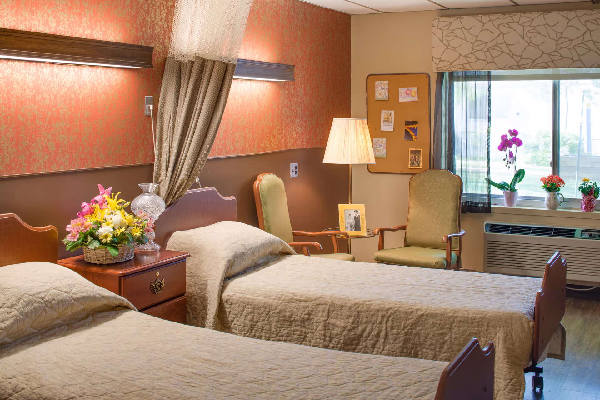 A cozy room with two single beds covered in beige quilts, a wooden nightstand with a basket of colorful flowers and a glass lamp, two green upholstered chairs, a floor lamp, a corkboard with pinned pictures, and a window with potted plants on the windowsill.