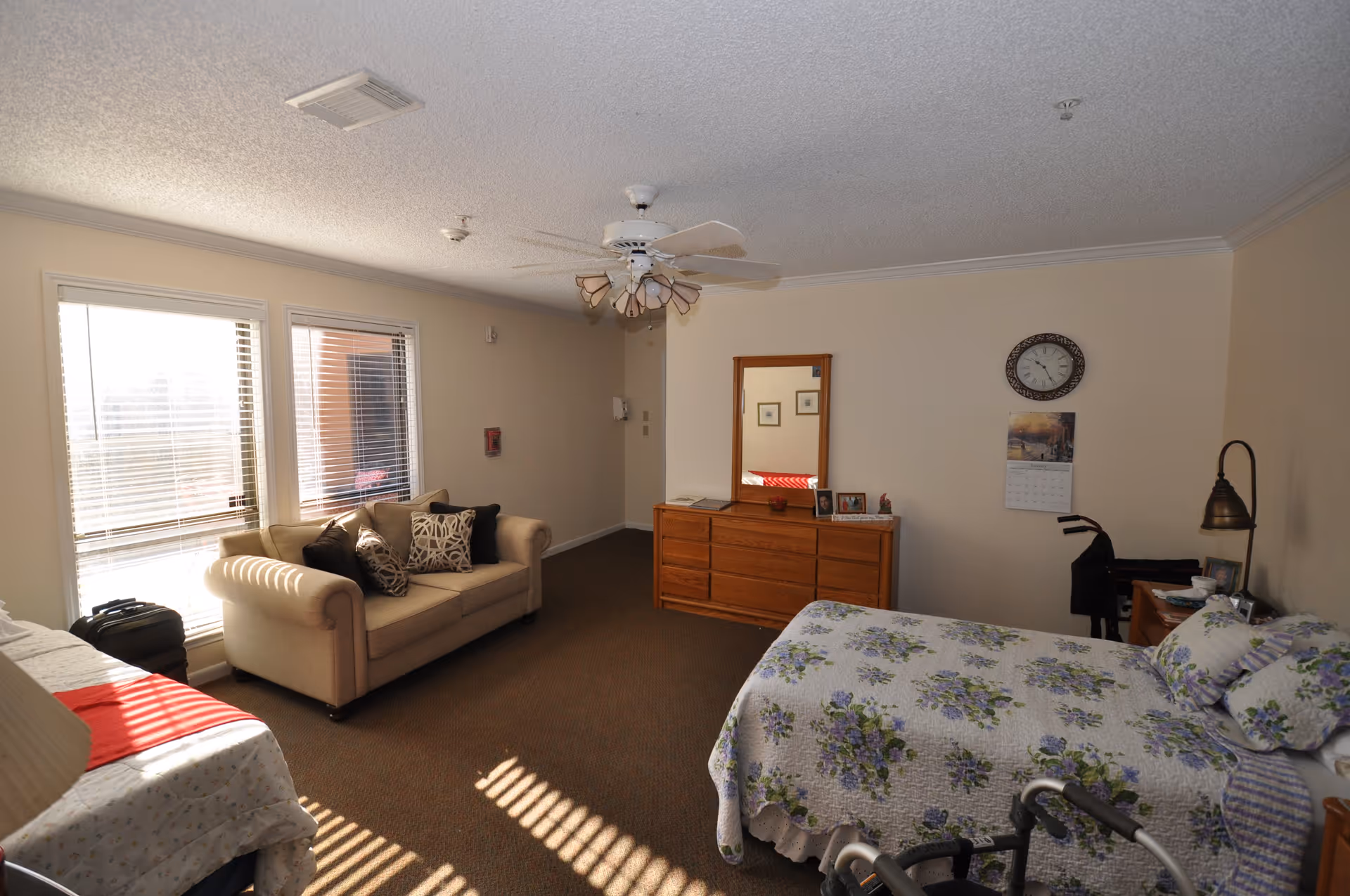 A well-lit bedroom in a senior living facility featuring a bed with floral bedding, a beige sofa with decorative pillows, a wooden dresser with a mirror, a wall clock, a calendar, and a walker near the bed. The room has large windows with blinds allowing natural light to enter, a ceiling fan with lights, and neutral-colored walls and carpet.