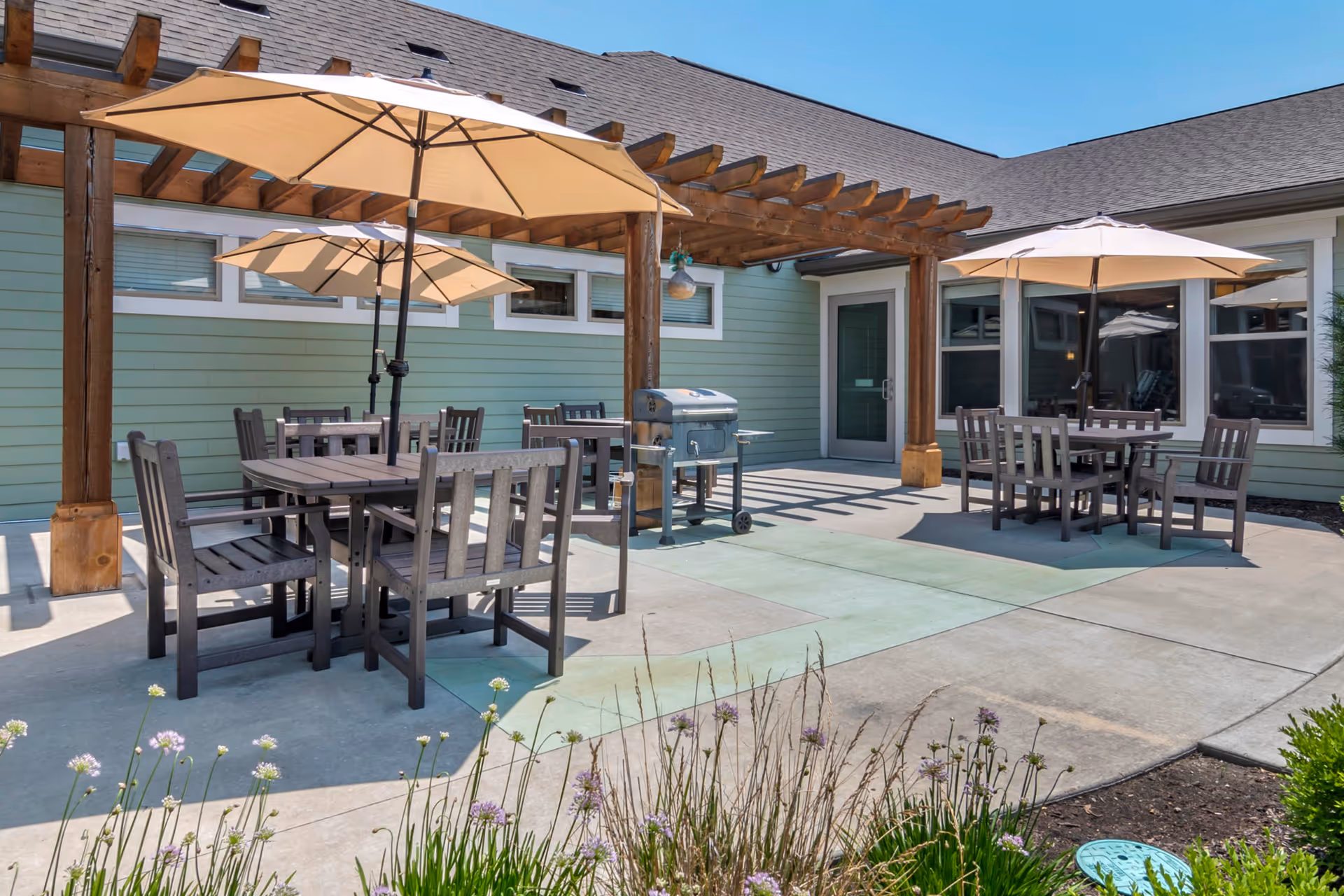 Sunlit outdoor patio with wooden tables and umbrellas, a pergola, and a grill in front of a light-green building.