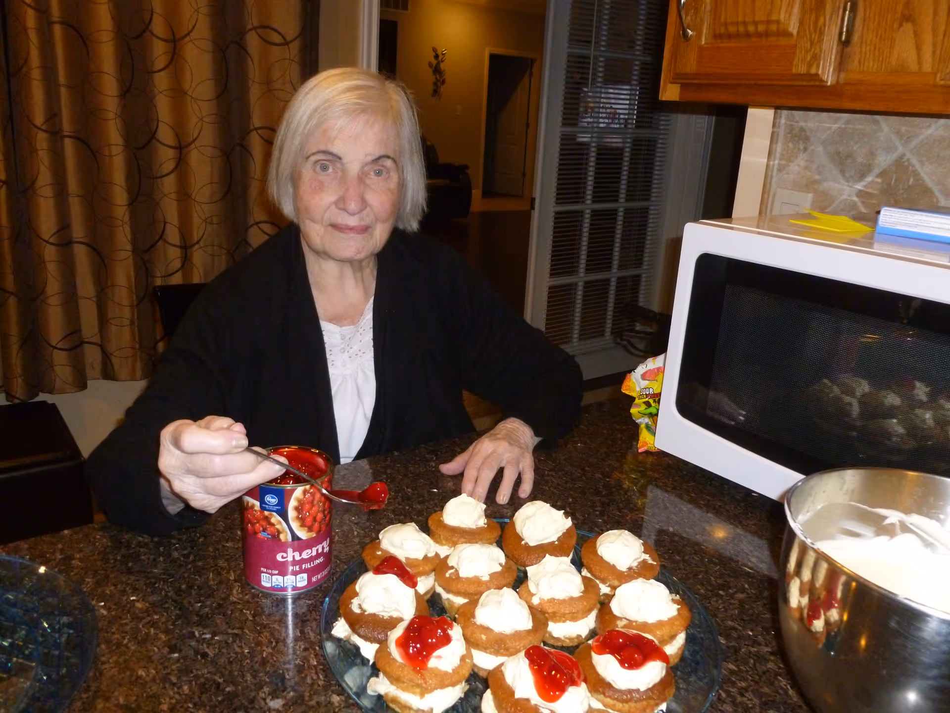 An elderly woman sitting at a kitchen counter, holding a spoon with cherry pie filling over a plate of small dessert cakes topped with whipped cream and cherry pie filling. A can of cherry pie filling, a mixing bowl with whipped cream, and a microwave are visible on the counter.