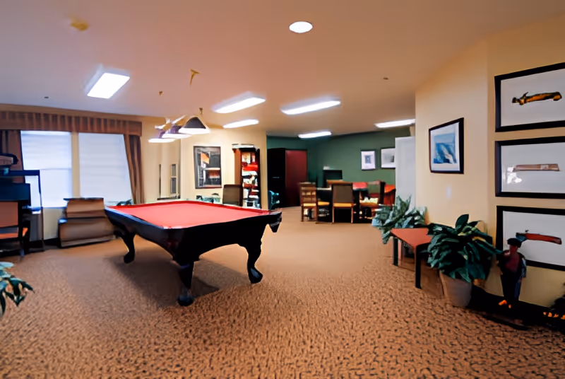 A spacious recreational room with a red felt pool table in the center, surrounded by comfortable seating including chairs and a bench. The room has beige walls adorned with framed artwork and plants placed near the walls. In the background, there is a dining or meeting area with a table and chairs. The room is well-lit with ceiling lights and has carpeted flooring.