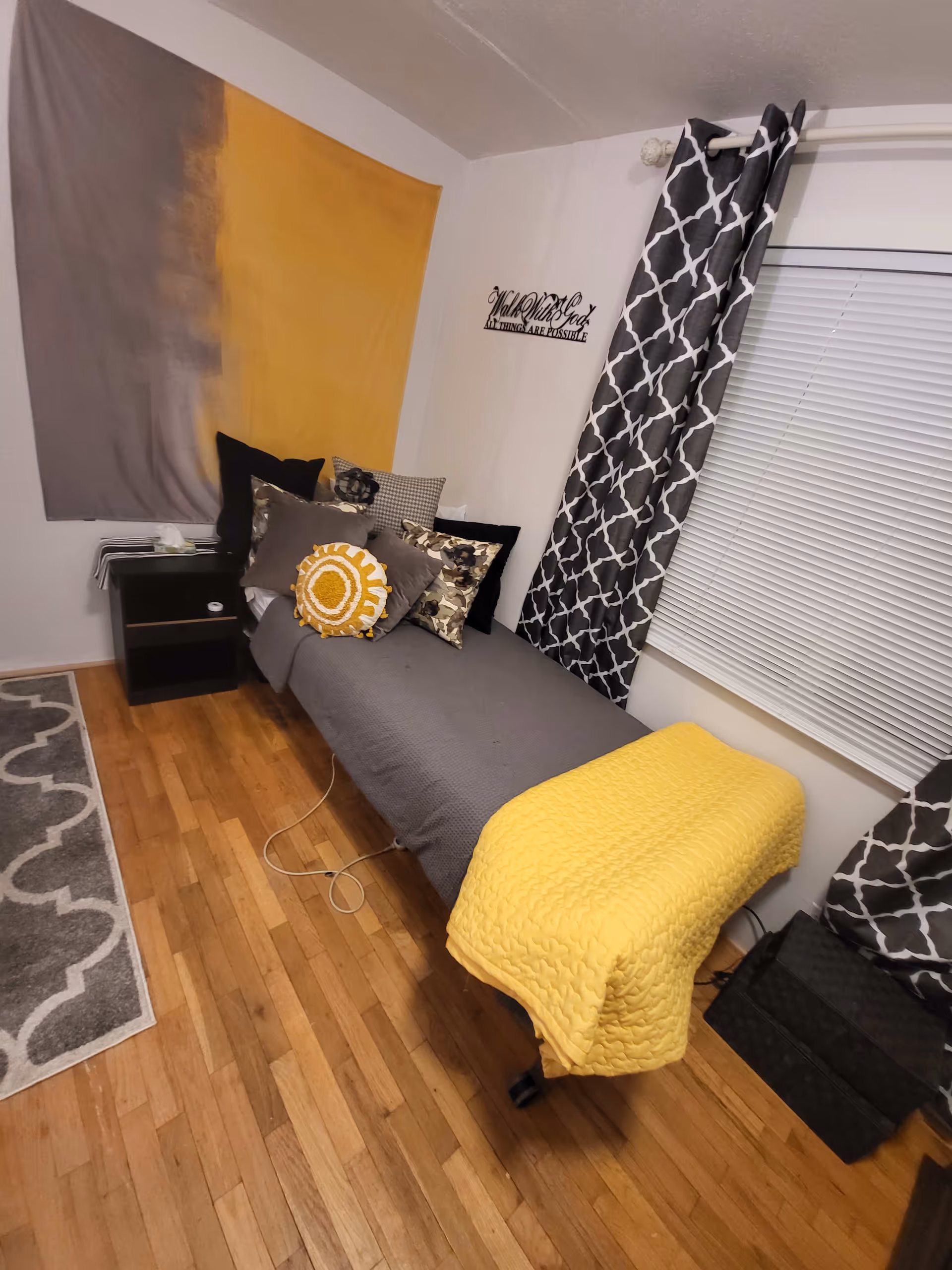 Small bedroom with a single bed dressed in gray and yellow bedding, decorative pillows, a nightstand, patterned curtains, and hardwood floor.