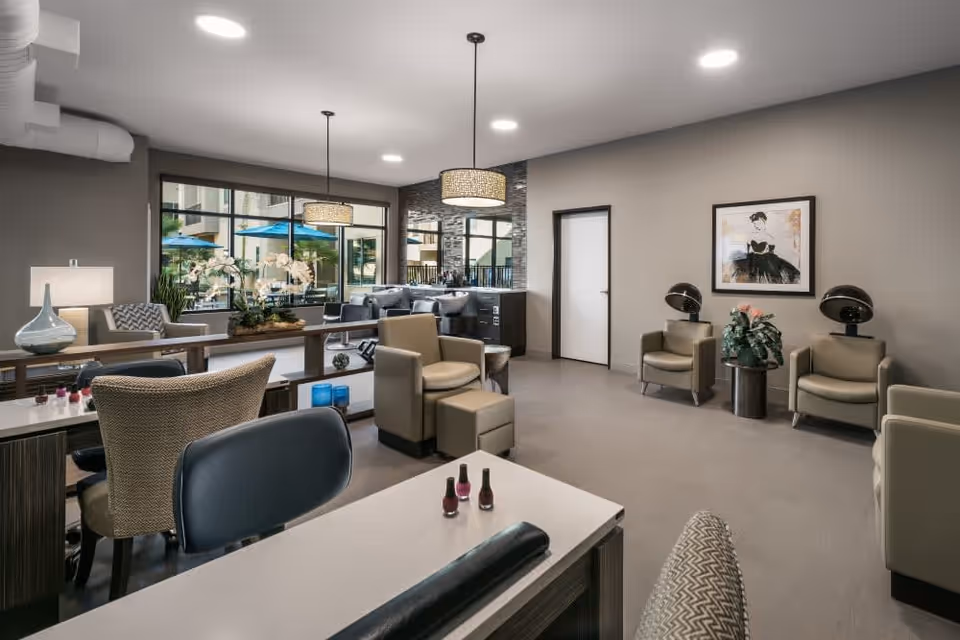 A modern and spacious salon area in a senior living facility with multiple salon chairs, manicure tables with nail polish bottles, comfortable armchairs, two hair drying stations, decorative plants, and large windows showing an outdoor patio with blue umbrellas.