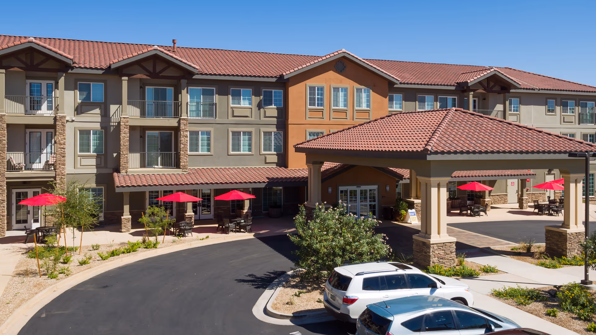 Exterior view of The Enclave at Gilbert Senior Living facility showing a three-story building with balconies, a covered entrance with a tiled roof, outdoor seating areas with red umbrellas, and parked cars in front under a clear blue sky.