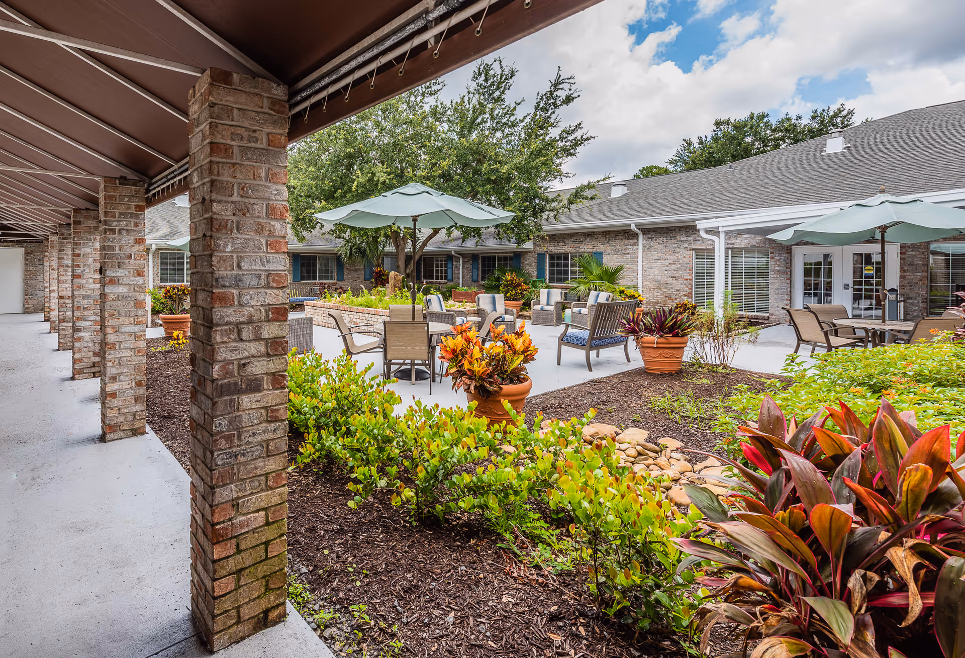 Outdoor courtyard area at The Colonnade at Carrollwood featuring a covered walkway with brick columns, patio seating with tables and umbrellas, various potted plants, shrubs, and trees under a partly cloudy sky.