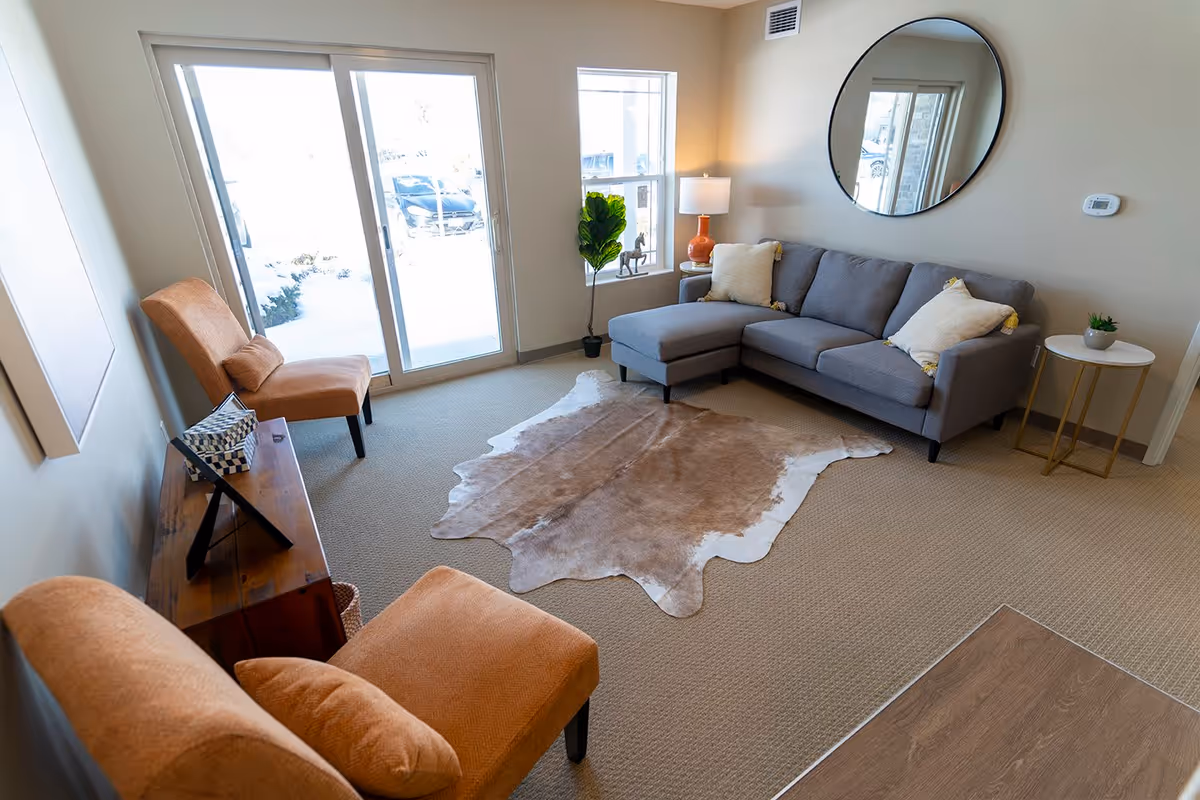 Bright living room featuring a gray L-shaped sectional beneath a round mirror, two orange chairs, a cowhide rug and sliding glass doors.