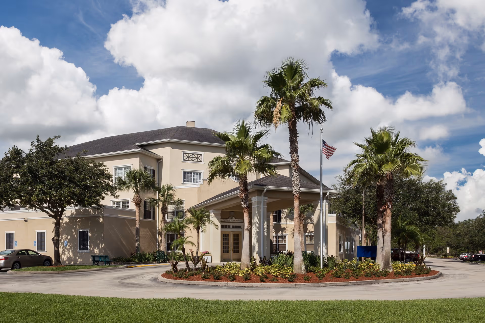 Exterior view of a senior living facility building with a circular driveway, palm trees, and an American flag on a flagpole under a partly cloudy sky.