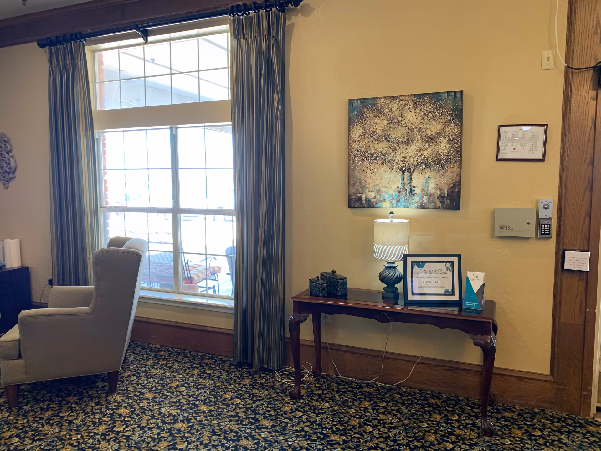 A cozy corner of a senior living facility featuring a large window with striped curtains, a comfortable armchair, and a wooden console table. On the table are a decorative lamp, framed certificate, and a small brochure. Above the table hangs a painting of a tree with golden leaves. The floor is covered with a patterned carpet.