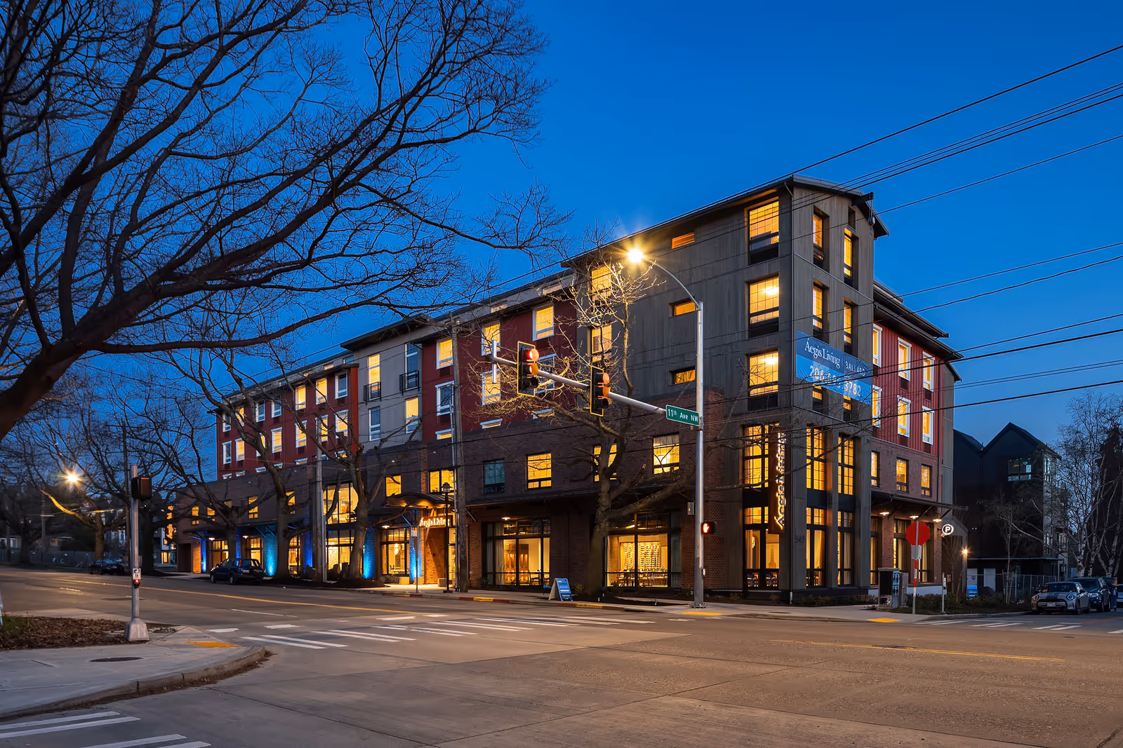 Exterior view of a multi-story senior living facility named Aegis Living Ballard at dusk, with warm lights glowing from the windows. The building is located at a street intersection with traffic lights and leafless trees in the foreground.