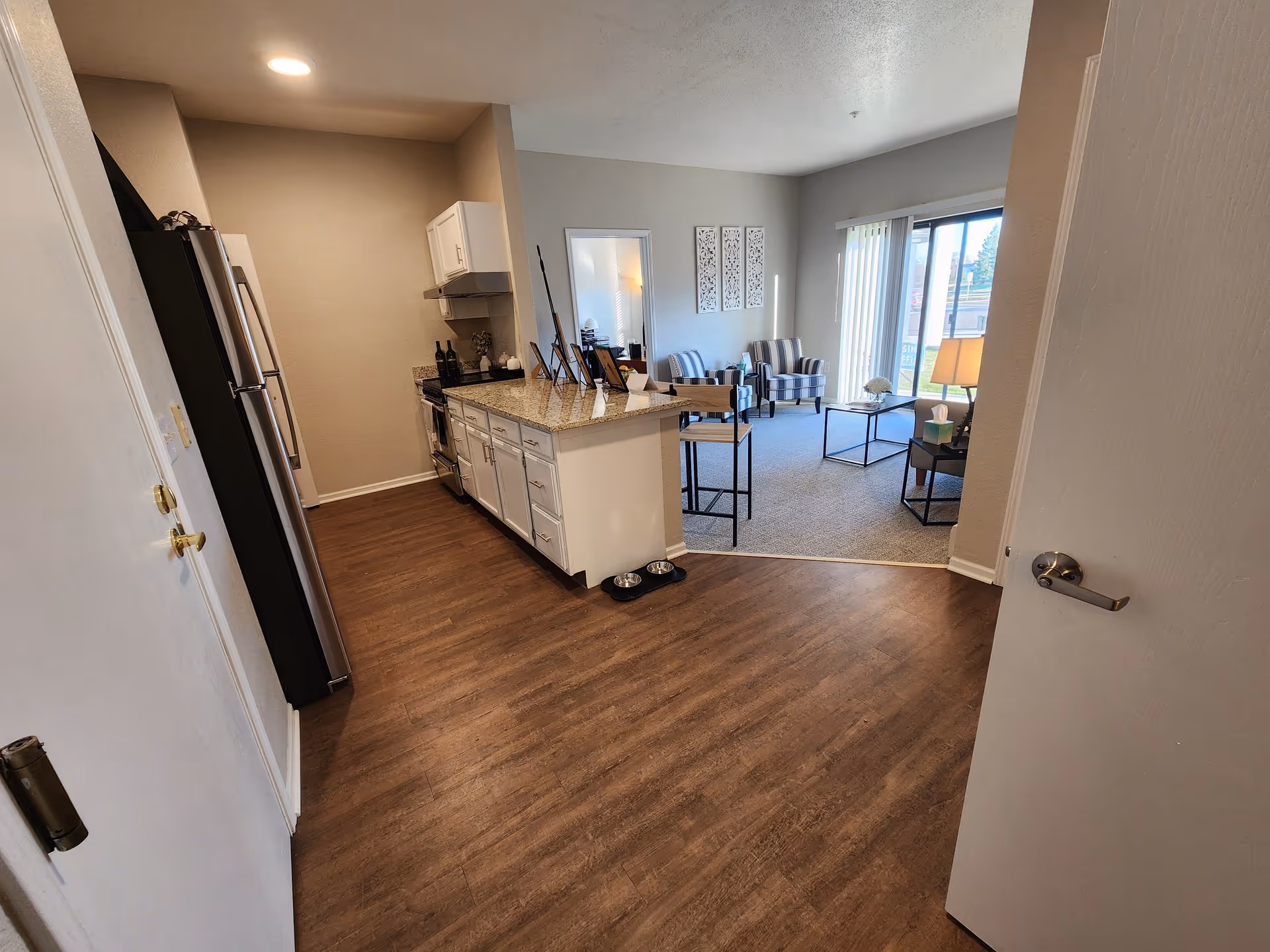 View of a modern kitchen area with wooden flooring and white cabinets, leading into a living room with carpeted floor, two striped armchairs, a coffee table, a side table with a lamp, and large sliding glass doors with vertical blinds letting in natural light.
