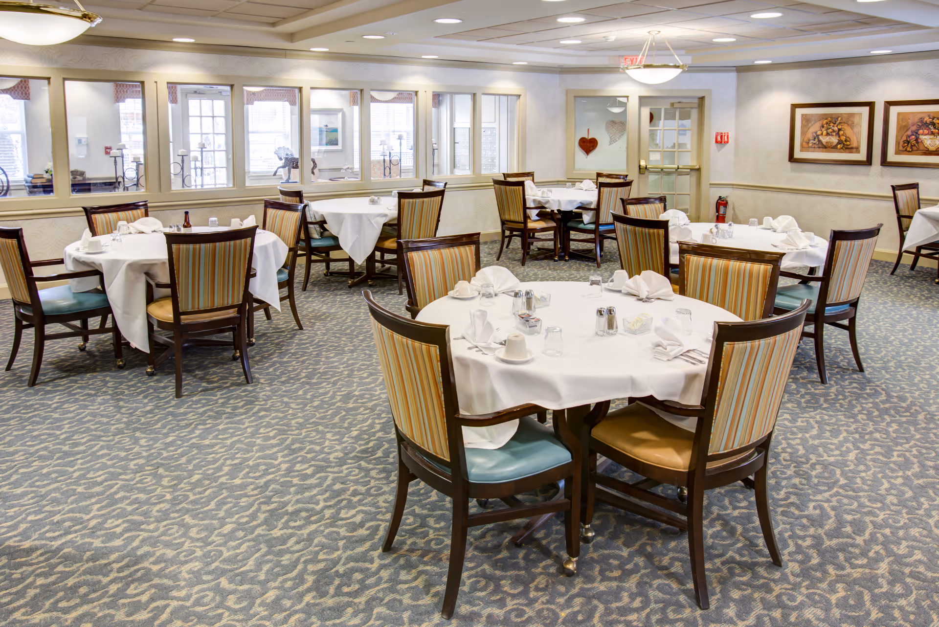 A dining room in a senior living facility with round tables covered in white tablecloths, each set with napkins, cups, glasses, and condiments. The room has patterned carpet, striped upholstered chairs, framed artwork on the walls, and windows letting in natural light.