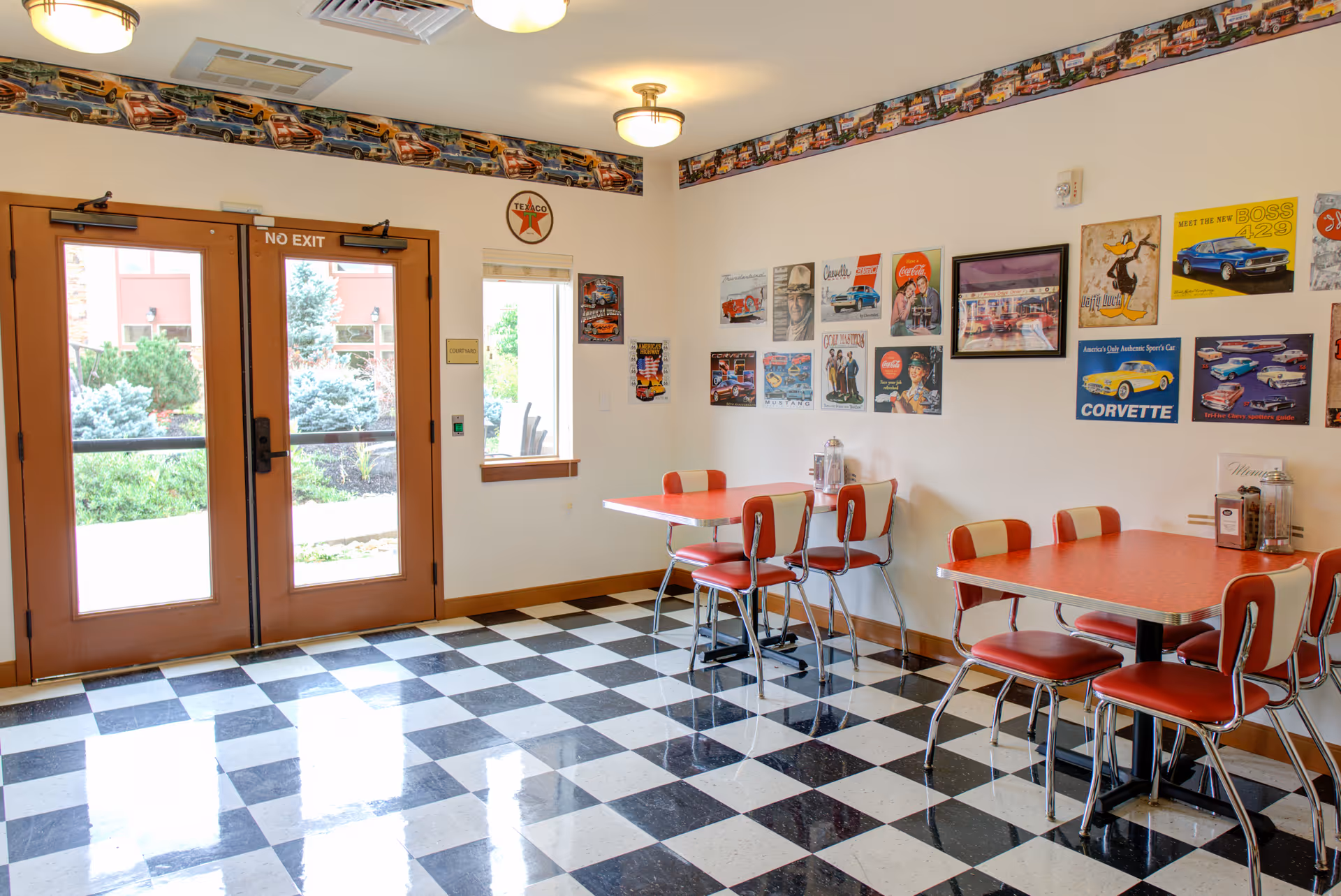 A retro-style dining area with black and white checkered floor tiles, red and white chairs, and red tables. The walls are decorated with vintage car and Coca-Cola posters, and there is a border near the ceiling featuring classic cars. Double glass doors lead outside to a courtyard with greenery visible through the windows.