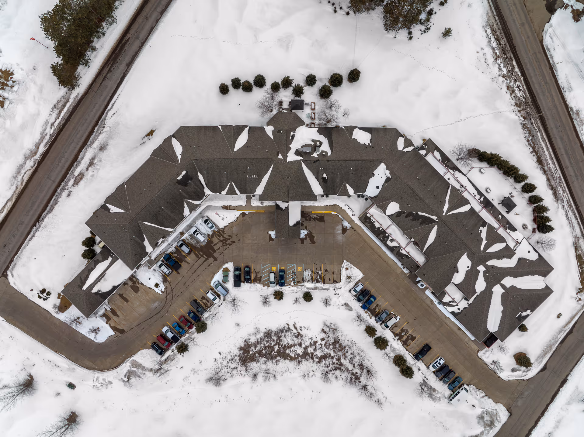 Aerial view of Sterling Pointe Senior Living facility covered in snow, showing the building's roof, surrounding parking lot with cars, and snow-covered grounds.