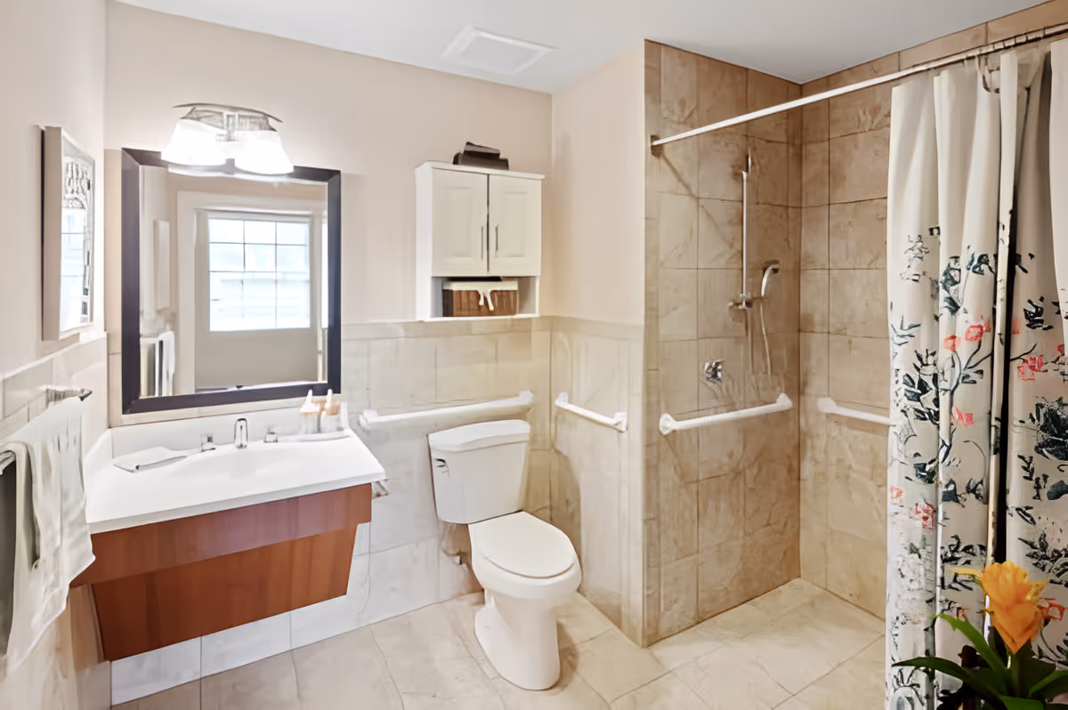 A clean and modern bathroom with beige tiled walls and floor. It features a white toilet, a wall-mounted sink with a wooden cabinet underneath, a large mirror with lights above it, and a walk-in shower with a floral-patterned shower curtain. There are grab bars installed near the toilet and inside the shower for accessibility. A small cabinet is mounted on the wall above the toilet, and a towel hangs on a rack beside the sink.