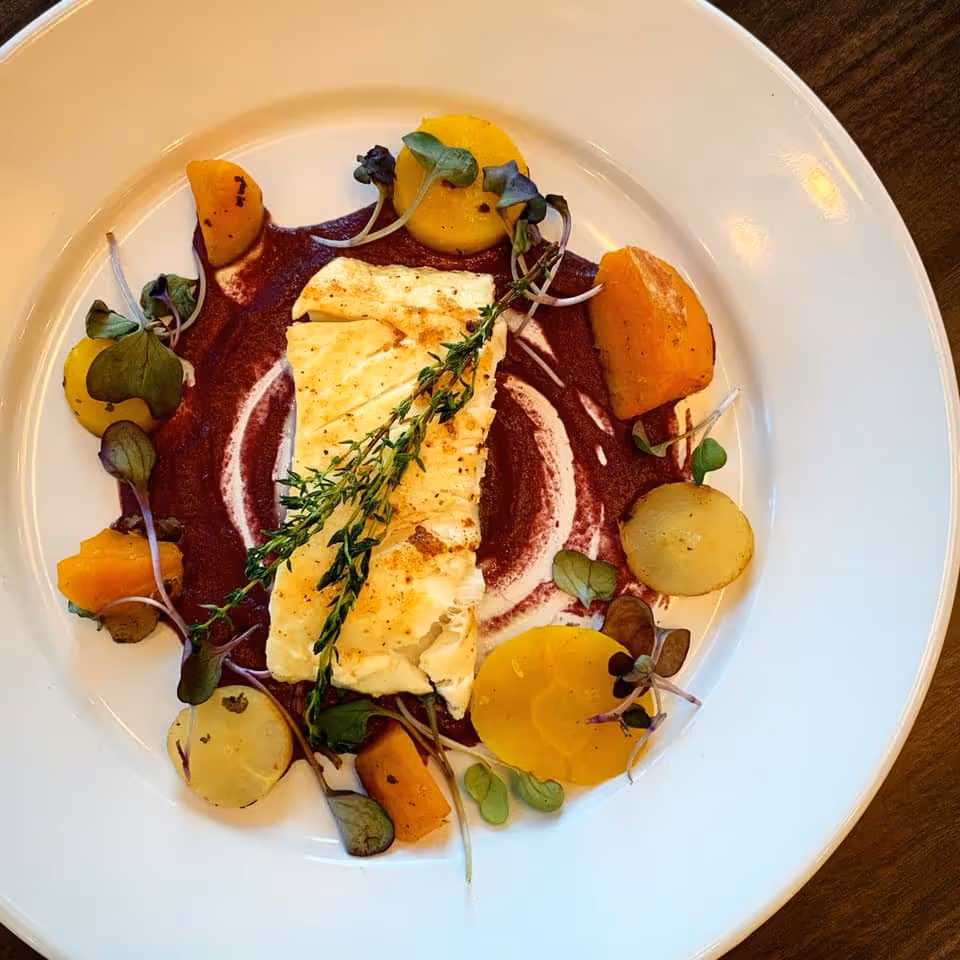 A plated gourmet dish featuring a seared white fish fillet on a purple sauce, garnished with thyme, microgreens and sliced root vegetables.