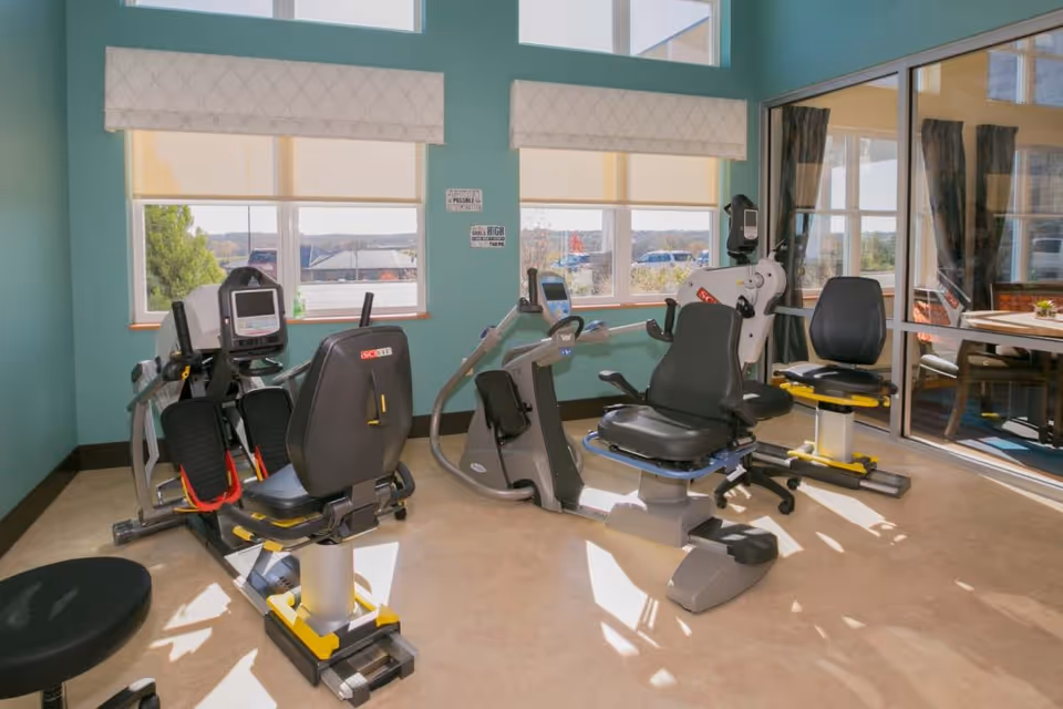 A bright fitness room with several recumbent exercise bikes and large windows overlooking the outside.