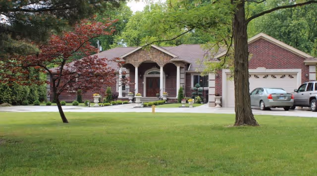 A single-story brick house with a front porch supported by columns, a well-maintained lawn, trees, and a driveway with parked cars.
