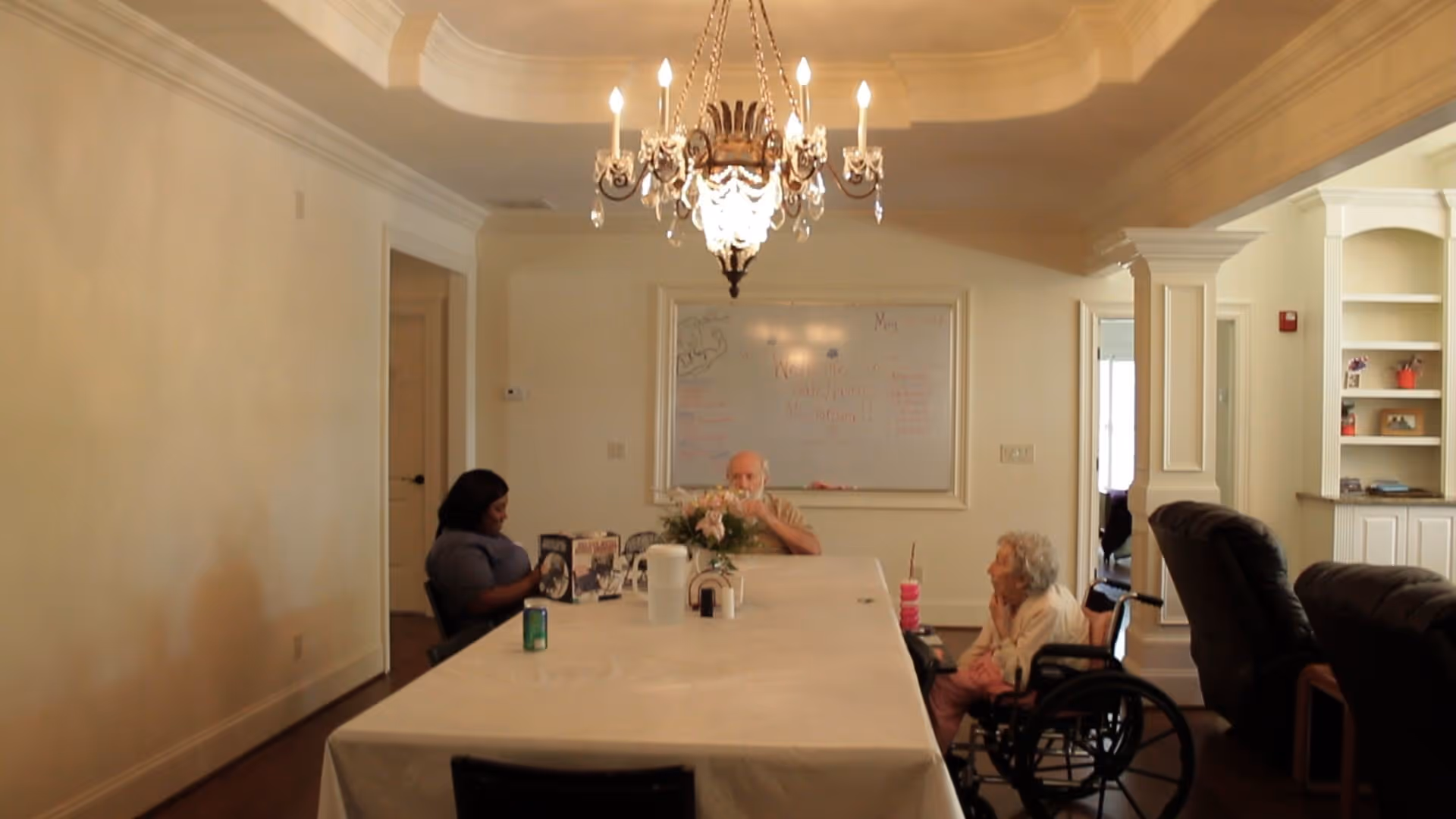 Communal dining room with a long table under a chandelier where residents and a caregiver sit, including an elderly woman in a wheelchair.