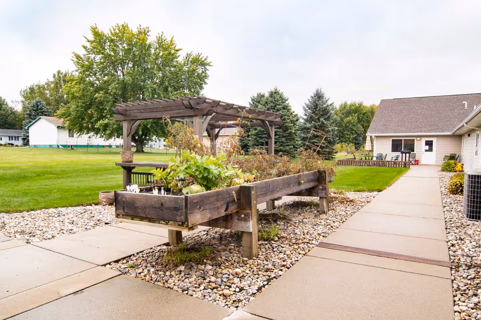 Outdoor garden area at Edgewood Flandreau featuring a raised wooden planter box with various plants, a wooden pergola with a bench underneath, a concrete walkway, and green lawn surrounded by trees and residential buildings in the background.