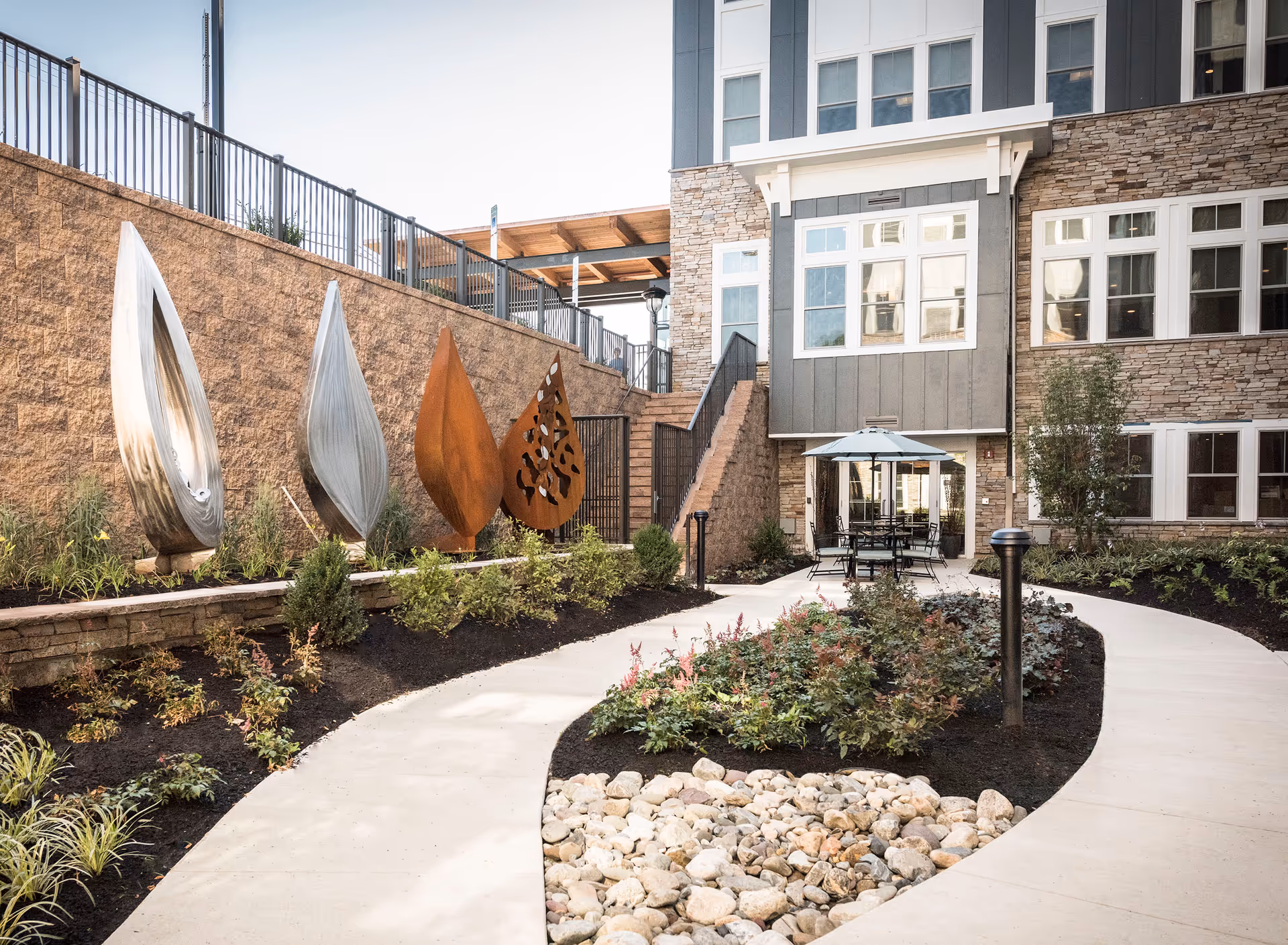 Outdoor courtyard area at Daylesford Crossing featuring a curved concrete pathway, landscaped garden beds with various plants and rocks, four large leaf-shaped sculptures made of metal, and a seating area with tables and umbrellas near the building entrance.