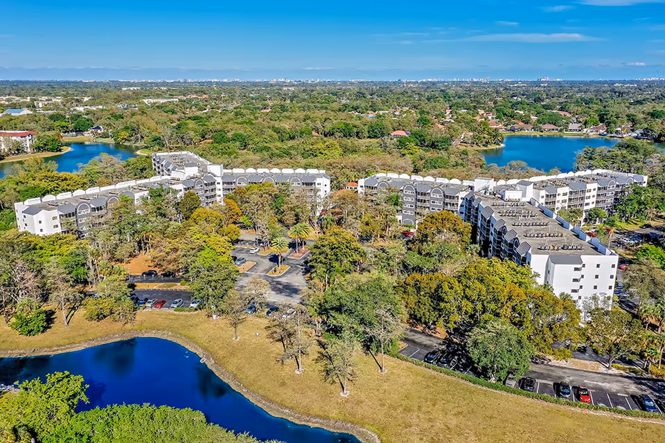 Aerial view of Forest Trace Senior Living facility surrounded by trees, parking lots, and several small lakes. The multi-story buildings are white with dark roofs, set in a lush green landscape under a clear blue sky.