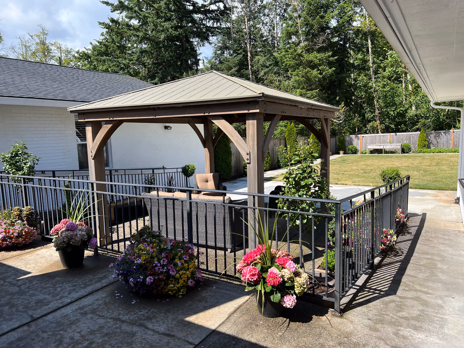 Outdoor patio area with a wooden gazebo featuring a metal roof, surrounded by black metal railings and several colorful flower pots. In the background, there is a grassy lawn, trees, and a white building with a window.