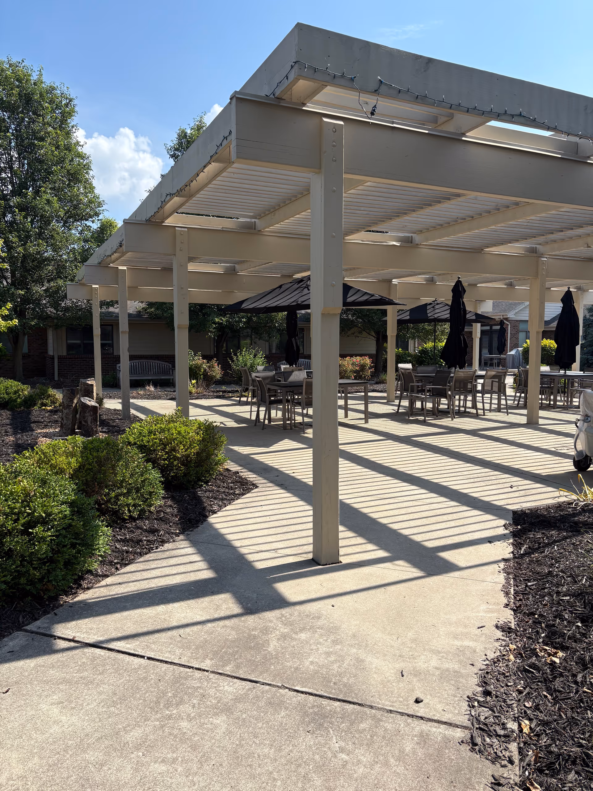 Outdoor senior living courtyard with a large pergola casting striped shadows over patio tables and chairs surrounded by landscaping.
